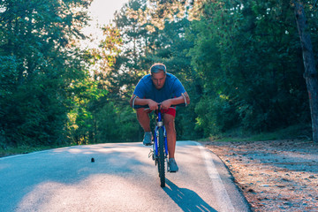 Older cyclist in his mid-40s riding a bike on a road through the woods and looking extremely tired.
