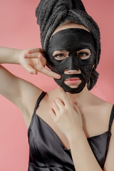 Young woman apply black cosmetic fabric facial mask on pink background. Face peeling mask with charcoal, spa beauty treatment, skincare, cosmetology. Close up