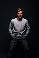 Fototapeta na wymiar Informally ( casual ) dressed blonde young man with sharp jawline in his 20's posing in a studio in front of a black background while wearing a white sweater.