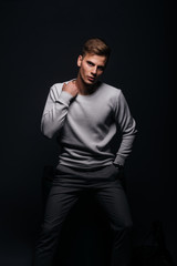 Obraz na płótnie Canvas Informally ( casual ) dressed blonde young man with sharp jawline in his 20's posing in a studio in front of a black background while wearing a white sweater.