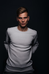 Informally ( casual ) dressed blonde young man with sharp jawline in his 20's posing in a studio in front of a black background while wearing a white sweater.