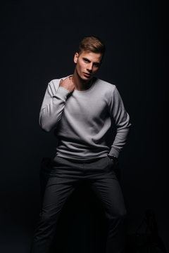 Informally ( casual ) dressed blonde young man with sharp jawline in his 20's posing in a studio in front of a black background while wearing a white sweater.