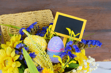 Detail of a spring time basket full of yellow and purple flowers flowers with a wooden background. Sparkly Easter eggs and a message board is added.