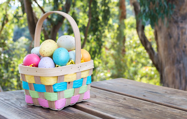 a wicker Easter basket filled with speckled Easter eggs sitting on a wooden picnic table overlooking a lush forest with copy space