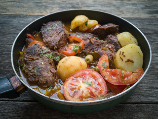 Meat with broth and vegetables in frying pan isolated on rustic wooden background.