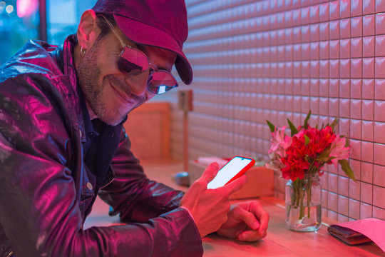 Young latin man with black cap, sun glasses and leather jacket looking his smartphone in a place with red neon lights - Powered by Adobe