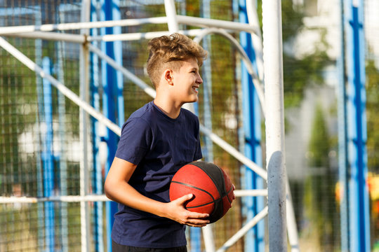 Cute smiling boy in blue t shirt plays basketball on city playground. Active teen enjoying outdoor game with orange ball. Hobby, active lifestyle, sport for kids.