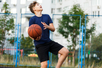 Cute smiling boy in blue t shirt plays basketball on city playground. Active teen enjoying outdoor game with orange ball. Hobby, active lifestyle, sport for kids.