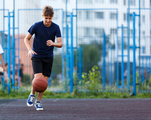Fototapeta premium Cute smiling boy in blue t shirt plays basketball on city playground. Active teen enjoying outdoor game with orange ball. Hobby, active lifestyle, sport for kids.