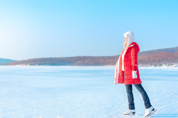 Young beautiful woman in red coat and figure skates stands on the frozen sea early in the morning. Winter outdoor activities concept. Leisure and lifestyle. Background with copy space.
