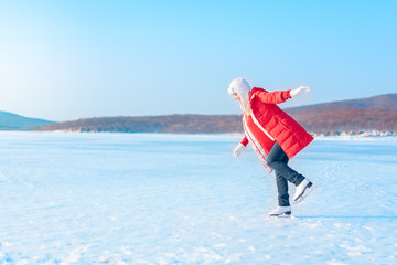 Young beautiful woman in red coat and figure skates stands on the frozen sea early in the morning....