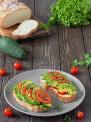Two ciabatta toasts with chopped avocado, tomato, olive oil, cheese, lettuce and black sesame seeds. Healthy vegetarian breakfast on a dark wooden table.