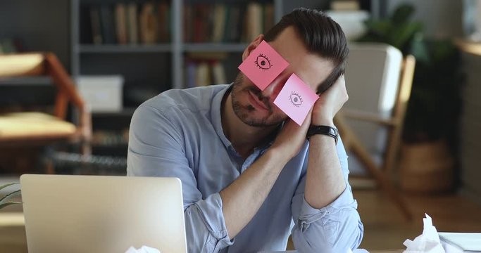 Funny lazy office worker napping at workplace covering eyes with sticky notes. Inefficient tired male employee pretends working sleeping with stickers on face sits at desk. Cheating to sleep concept