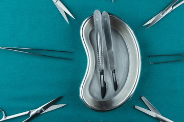 Surgical instrument, stainless steel scalpel handle number 3 with blade number 10 and scalpel handle number 4 with blade number 24 in emesis or plus basin and tweezers scissors on surgical green drape