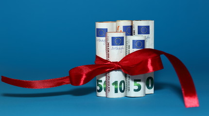 Banknotes of euro currency  rolled in rolls, tied with decorative red ribbon, bow isolated on blue  background, concept of economical / financial gift / present