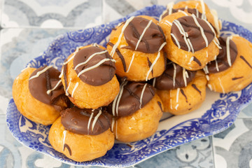 Profiteroles, coated in chocolate, stacked on a pattermed blue and white plate.  On a tile background