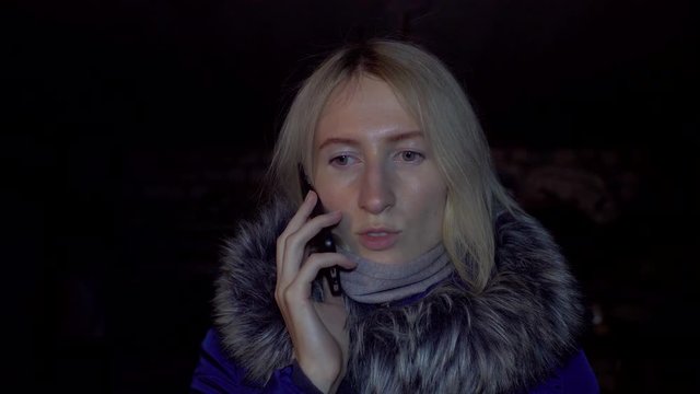 Portrait of shocked scared young woman. She is lit by police flashing lights and is talking on phone. woman is scared, desperately wants something, covering her mouth with hands of problem.