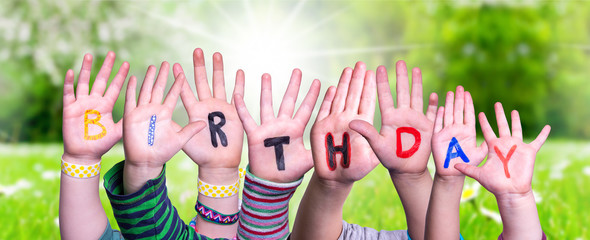 Children Hands Building Colorful Word Birthday. Sunny Green Grass Meadow As Background