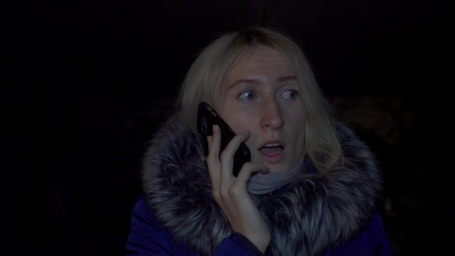 Portrait of shocked scared young woman. She is lit by police flashing lights and is talking on phone. woman is scared, desperately wants something, covering her mouth with hands of problem.