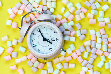 Top View of Pastel Shaped Marshmallow Candies and alarm clock with Some Scattered on the Pale yellowTable