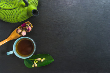 Obraz na płótnie Canvas Tea time composition. Green iron teapot, cup of tea, wooden spoon with rose buds and green tea leaf on black slate background. Space for text, top view