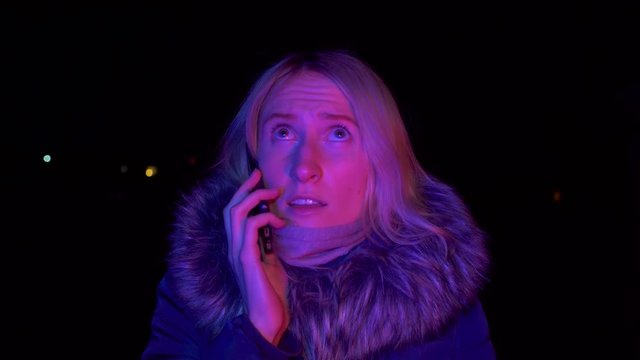 Portrait of shocked scared young woman. She is lit by police flashing lights and is talking on phone. woman is scared, desperately wants something, covering her mouth with hands of problem.