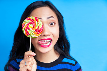 Close up of young asian woman sticking out tongue covering her eye with colorful lollipop