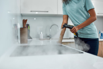 Service at work. Cropped view of a woman in uniform cleaning electric stove with steam cleaner while working in the modern kitchen