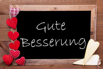 Balckboard With German Text Gute Besserung Means Get Well Soon. Red And Yellow Heart Decoration. Brown Wooden Background