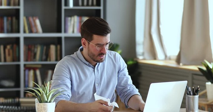 Ill male worker sitting at workplace blowing nose in tissue. Sick unhealthy businessman feeling fever having flu cold symptoms at work. Virus infection seasonal diseases in office environment concept