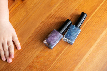 two shiny nail polishes, blue and purple, lie on a wooden surface. child's hand reaches for bottles with varnish
