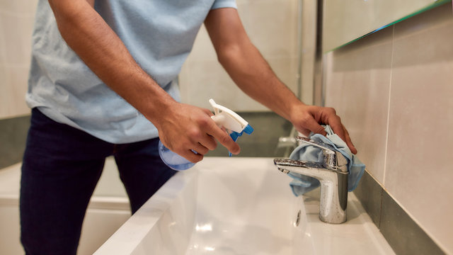 Perfectly clean. Cropped photo of male cleaner in uniform cleaning water tap with spray detergent and wipe cloth in the bathroom