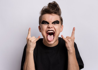 Crazy teen boy with spooking make-up making Rock Gesture on gray background. Teenager in style of punk goth dressed in black shows tongue and doing heavy metal rock sign. Problems of transitional age