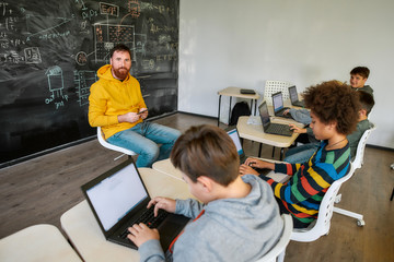 Where Students Are Achievers. Portrait of male teacher looking at the camera, while sitting with tablet pc near the blackboard during a lesson in modern smart school. Selective focus