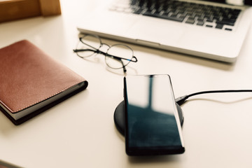 Smart phone being charged on wireless charger alongside with laptop, glasses, and notebook on table...