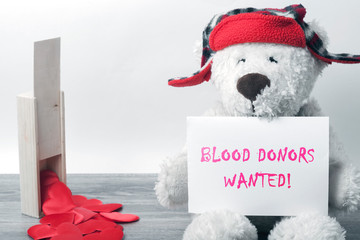 A white Teddy bear in a red hat sits on a table and holds a sheet of white paper with the...