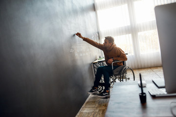 Obraz na płótnie Canvas Working on presentation. Young male professional designer in a wheelchair writing something on big blackboard in the modern office. Portrait of disabled young man sketching some ideas