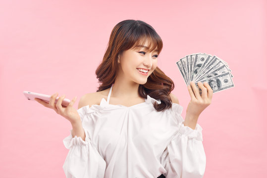 Image of a beautiful excited young woman posing isolated over pink wall background holding money and using mobile phone.
