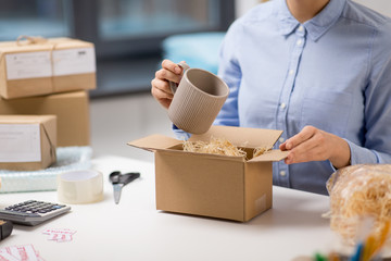 delivery, mail service, people and shipment concept - close up of woman packing mug to parcel box...