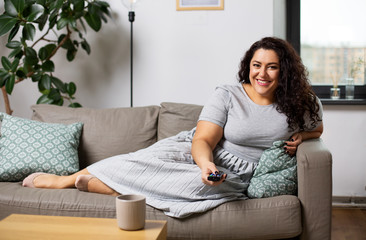 Fototapeta premium people and leisure concept - happy young woman lying on sofa with remote control and watching tv at home
