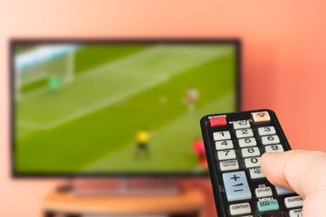 Caucasian man watching soccer or football on TV at home. Changing channels and adjusting volume with television remote control. 