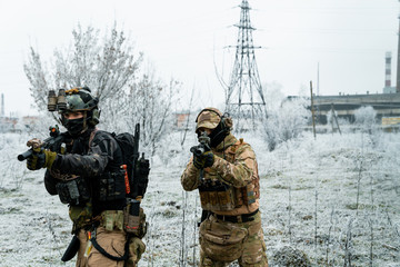 Men in camouflage cloth and black uniform with machineguns with factory on background. Soldiers with muchinegun aims aiming