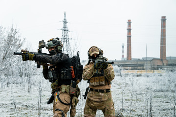 Naklejka premium Men in camouflage cloth and black uniform with machineguns with factory on background. Soldiers with muchinegun aims aiming. Horizontal photo.