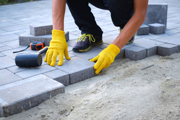 The master in yellow gloves lays paving stones in layers. Garden brick pathway paving by professional paver worker. Laying gray concrete paving slabs in house courtyard on sand foundation base.