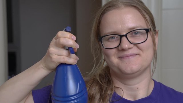 Young Caucasian woman with know-it-all face expression holds the plastic blue sprayer in hands. Sanitation of rooms and objects concept