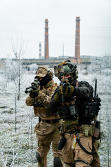 Men in camouflage cloth and black uniform with machineguns side to side with factory on background....