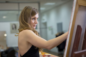 Fototapeta na wymiar Woman artist in art studio paints a picture on a canvas in easel, profile portrait.