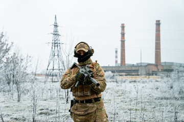 Naklejka premium Man in camouflage uniform hold machinegun. Soldier in the winter with factory on background. Horizontal photo side view.