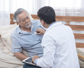 Fototapeta premium Asian senior elderly old man with mask on lying on sofa while young caucasian doctor sit on knee check his heart beat and old senior woman sit beside with care and worry .healthcare concept.
