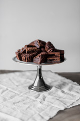 Pieces of chocolate brownie on a silver tart plate on white linen and wooden table